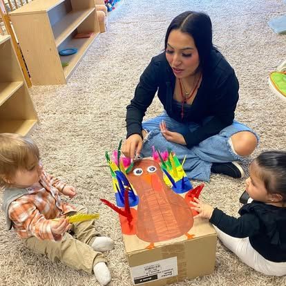 An adult and two toddlers play with a turkey craft made from a cardboard box and colorful feathers on a carpeted floor.