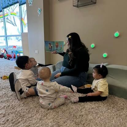 An adult sits on a carpeted floor reading a book to four children in a brightly lit room with wall-mounted lights.