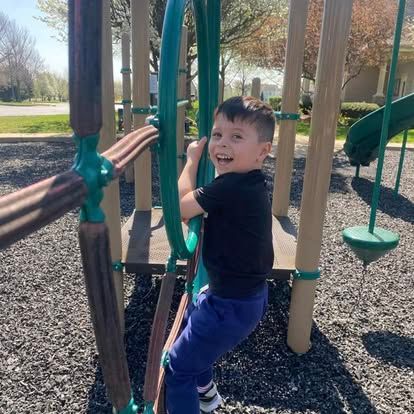 A child with a joyful expression climbs a green playground structure outdoors on a sunny day.