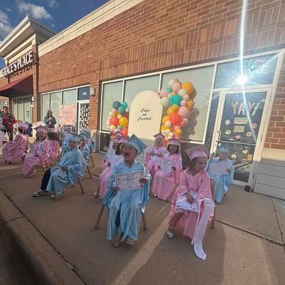 Children in graduation caps and gowns sit on chairs outside a building, holding certificates near balloon decorations.