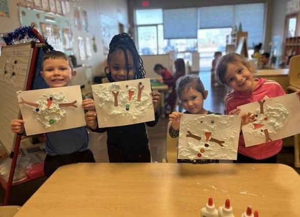 Four children stand at a table in a classroom, smiling as they display their snowman crafts made with white puff paint.