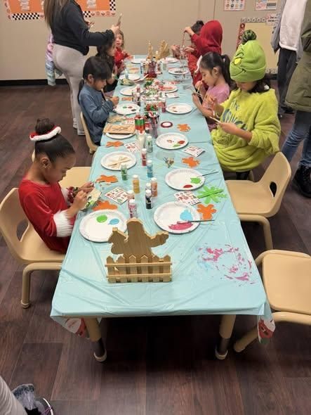 A group of children sits at a long table with blue paper, decorating holiday-themed crafts with paint and supplies.