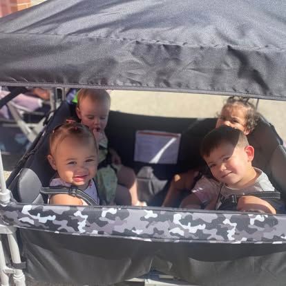 Four children sitting inside a camouflage-patterned stroller wagon with a black canopy outdoors.