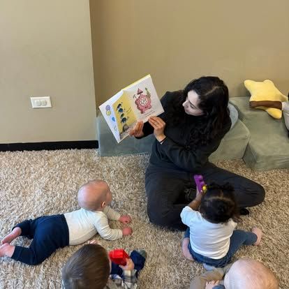 An adult sits on a carpeted floor reading a book to four children in a play area.