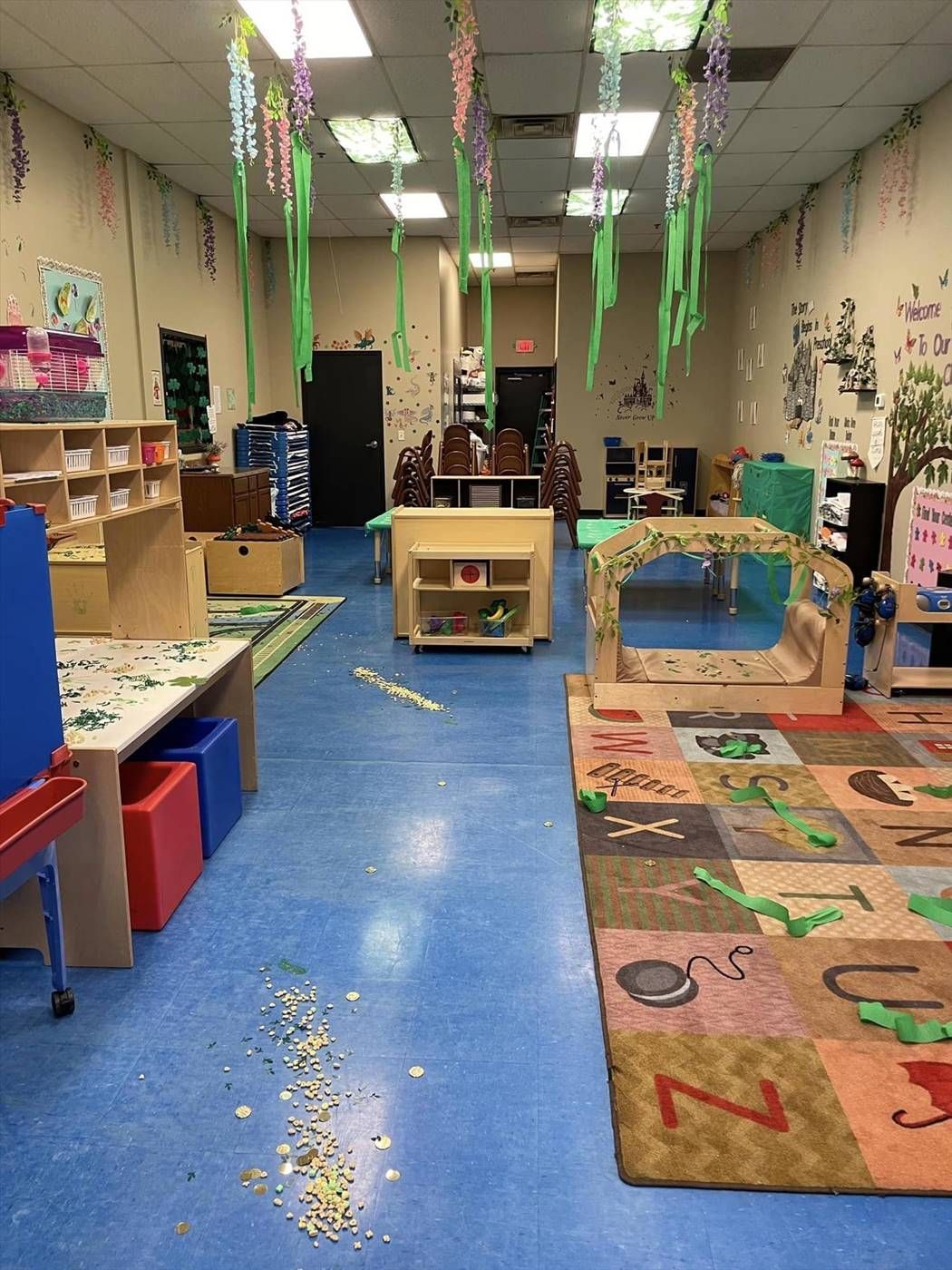 A classroom with blue flooring, play areas, wooden furniture, and hanging green decorations, with debris on the floor.