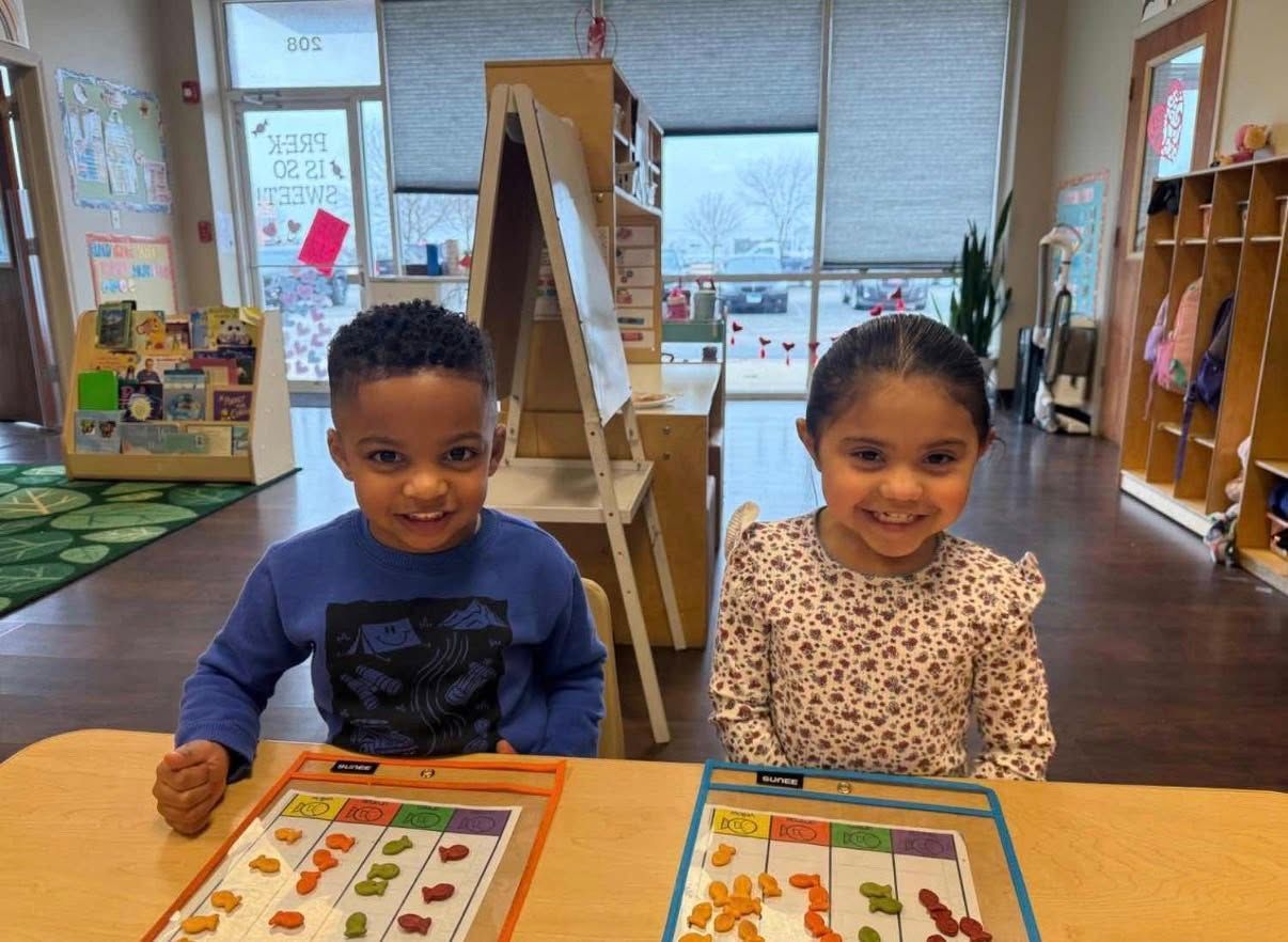 Two children smile while sorting colorful snacks into columns on activity sheets at a table in a classroom.