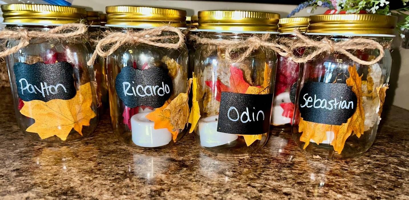Four glass jars with gold lids, filled with faux autumn leaves and small candles, labeled Payton, Ricardo, Odin, Sebastian.