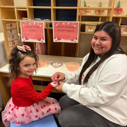 A teacher and child seated at a desk with heart-themed materials for a Valentine's craft activity.