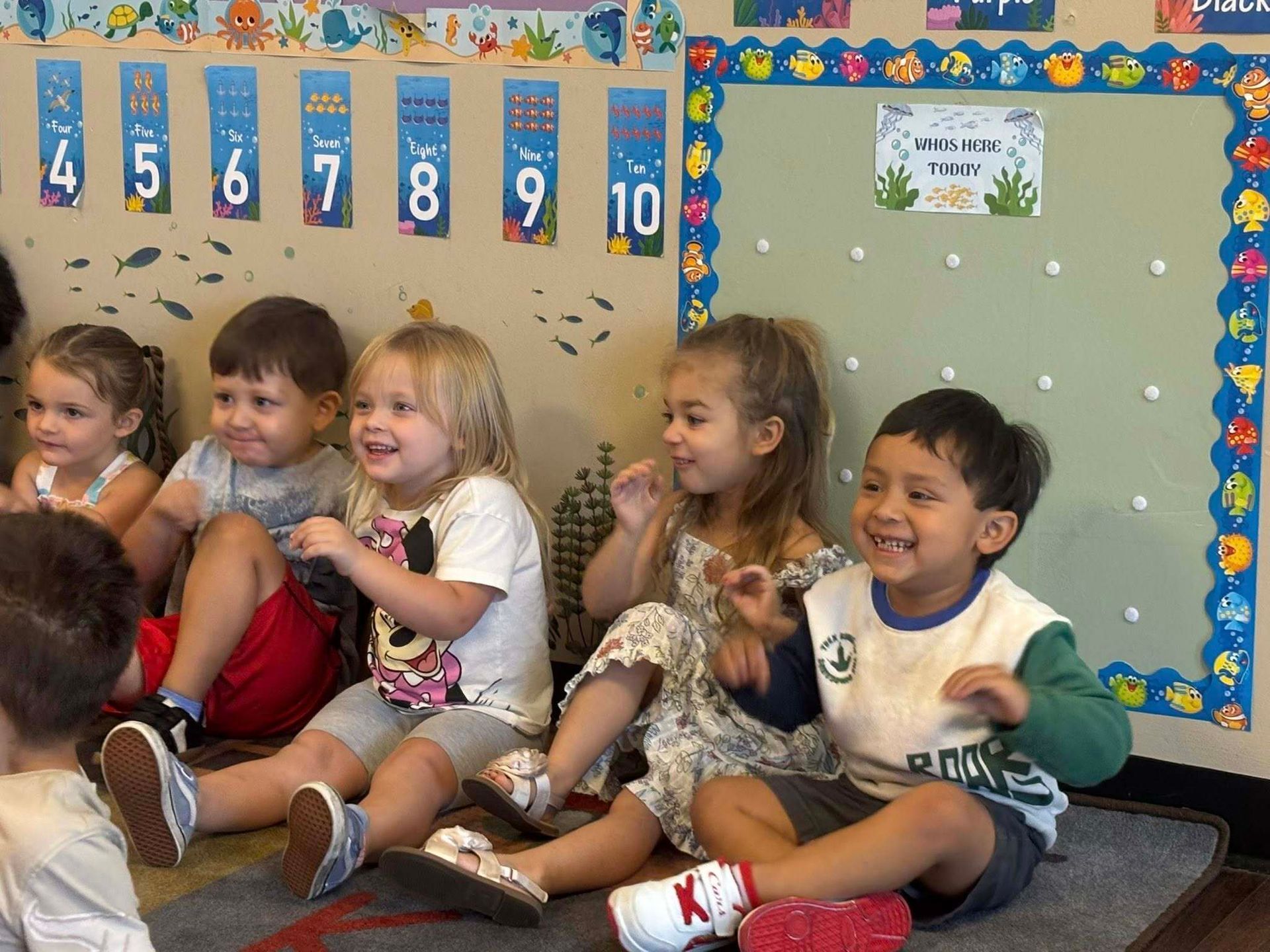 A group of young children sit on the floor in a classroom decorated with numbers and sea-themed borders, smiling.