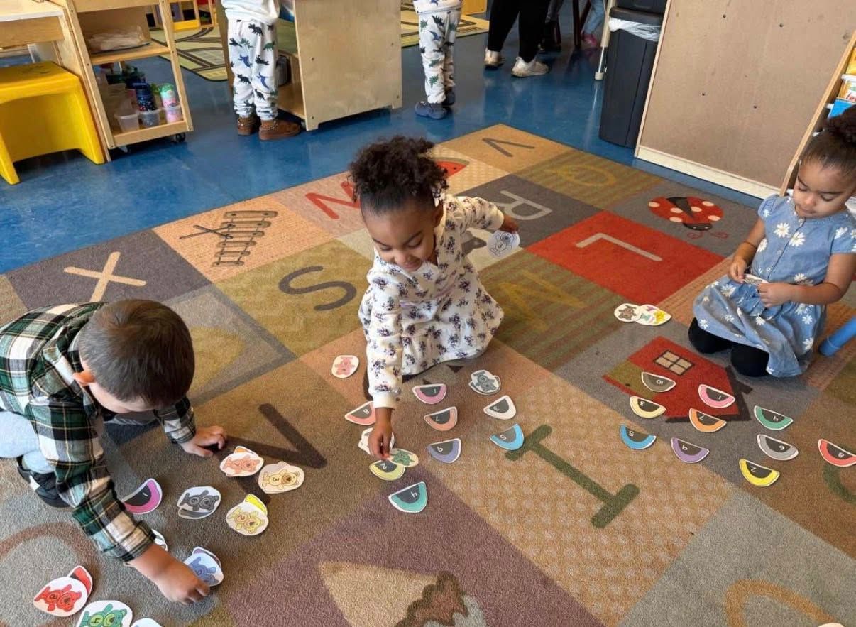 Three children sit on an alphabet-patterned rug, sorting small, colorful cards during a classroom activity.
