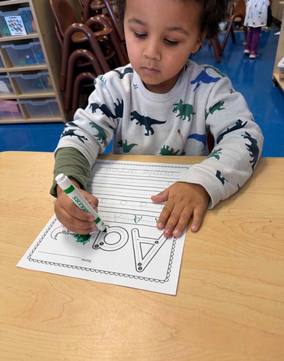 A child in a dinosaur-print sweater uses a green marker to trace the letters A and a on a classroom worksheet.