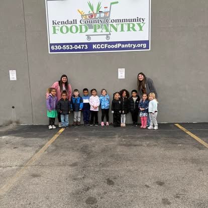 A group of children and two adults stand in front of a Kendall County Community Food Pantry sign on a gray wall.
