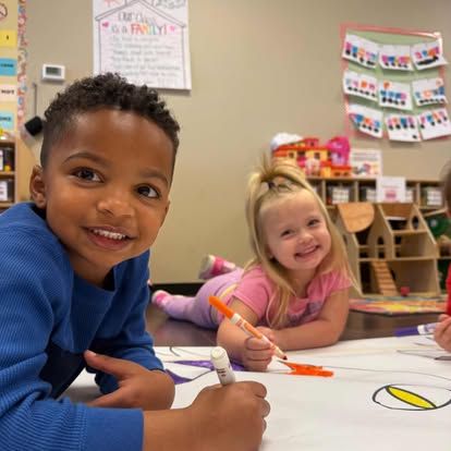 Two children lie on the floor in a classroom, smiling as they color together on a large sheet of paper.