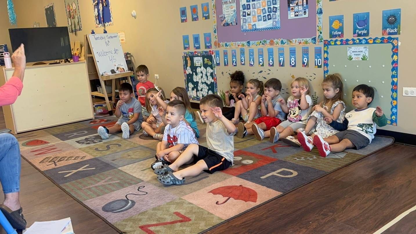 A group of children sit on a colorful alphabet rug in a classroom, looking toward a teacher during a lesson.