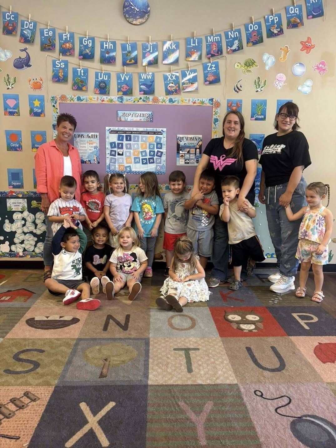 A group of children and teachers posed for a photo in a classroom with alphabet decor and an educational rug.
