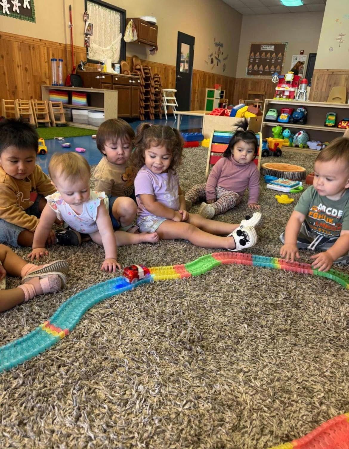 A group of young children plays with a colorful toy car track on a carpeted floor in a daycare room.