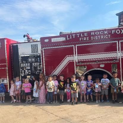 A group of children stand in front of a red Little Rock-Fox Fire District fire engine on a sunny day.