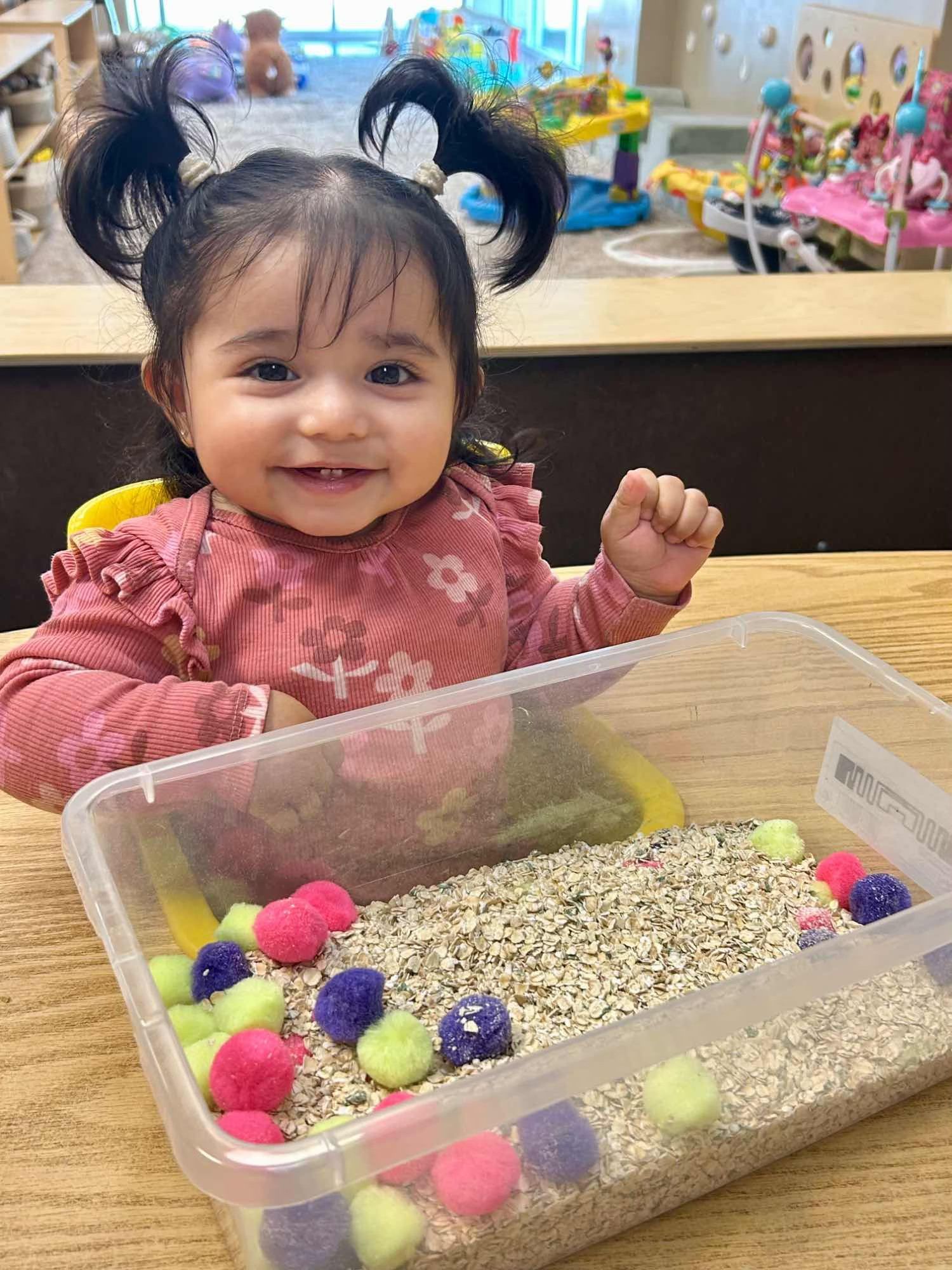 A happy child sits at a table with a sensory bin filled with oats and colorful pom-poms.