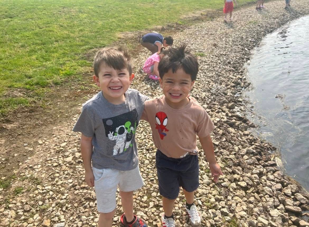 Two smiling children stand with their arms around each other on a rocky shoreline next to a body of water.