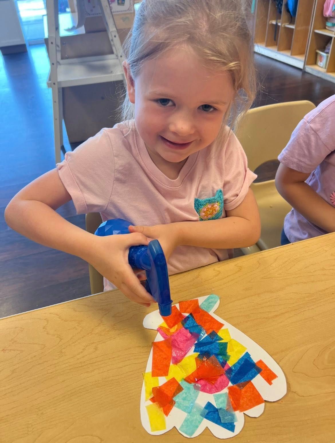 A smiling child uses a small blue spray bottle to wet colorful tissue paper shapes on a white paper cutout at a table.