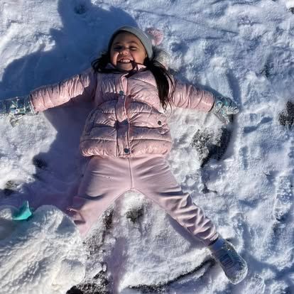 A smiling child in a pink coat, pants, and a grey hat lying in the snow to make a snow angel.