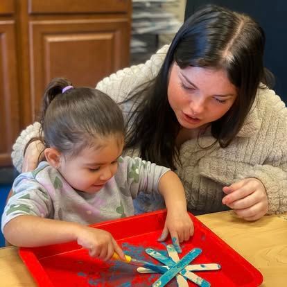 A person supervises a child painting blue star-shaped popsicle sticks on a red tray.