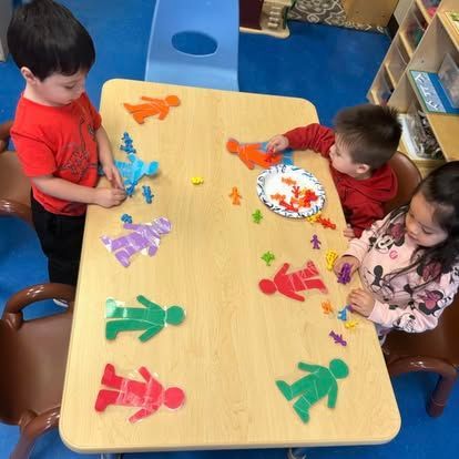 Three children sit at a table playing with colorful human-shaped plastic figures and puzzle pieces in a classroom.