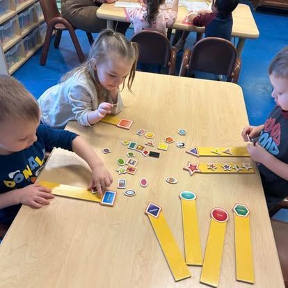 Children sit around a table sorting colorful shape cutouts onto corresponding wooden boards in a classroom setting.