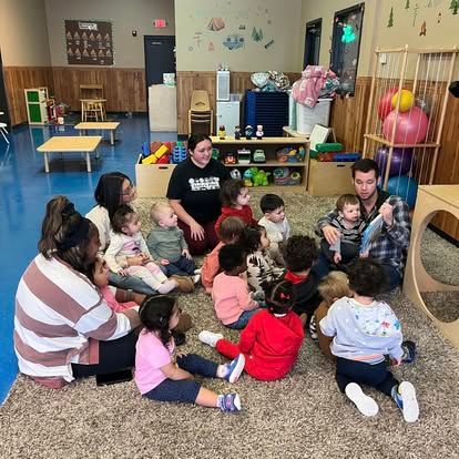 Two adults and a group of young children sit on a rug in a classroom, with one adult reading from a book to the group.