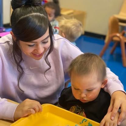 A smiling person in a light purple shirt guides a toddler playing with items on a yellow tray in a bright classroom.