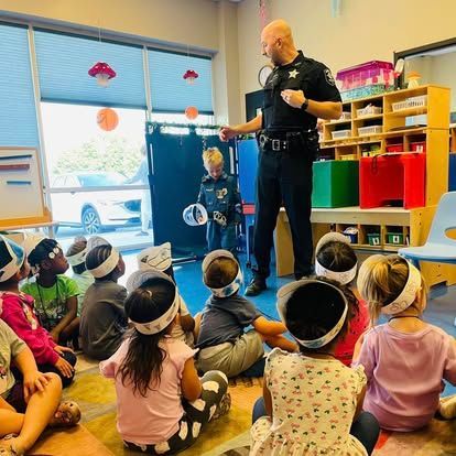 A police officer in uniform stands in a classroom, talking to a child while other children sit on the floor nearby.