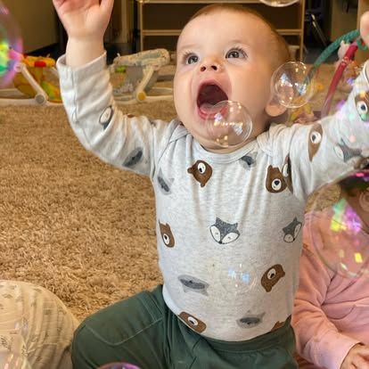 A baby wearing a gray animal-print shirt reaches up toward floating bubbles, eyes wide and mouth open in excitement.