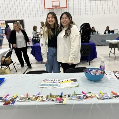 Two people stand smiling behind a table at an indoor event, with photos clipped to the front edge of the table.