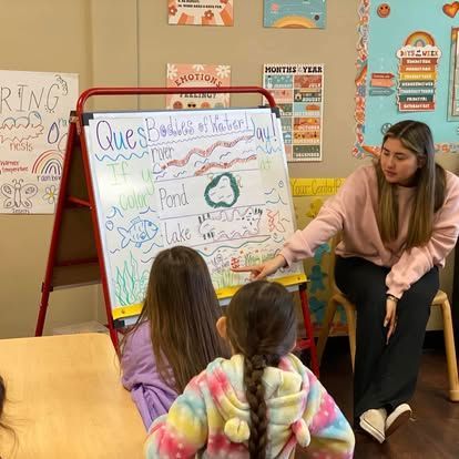 A teacher points to a whiteboard drawing about bodies of water while two students sit in front, facing the board.