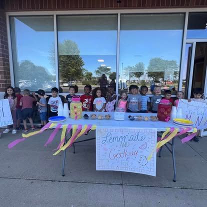 A large group of children stand behind a table labeled "Lemonade" with pitchers and cups outside a building.