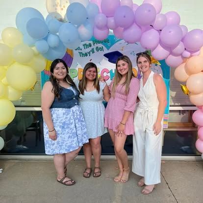 Four people stand smiling together in front of blue and silver balloon decorations in a classroom setting.