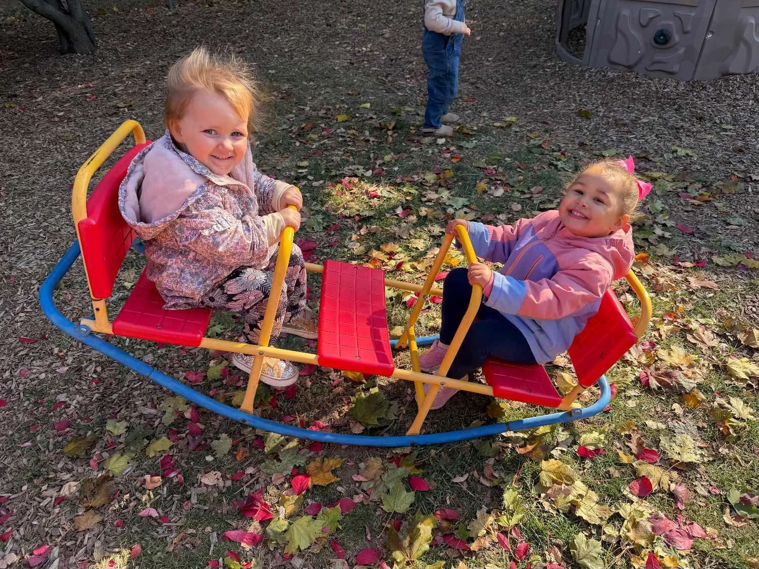 Two children smiling while playing on a red and yellow seesaw on a playground covered with fallen autumn leaves.