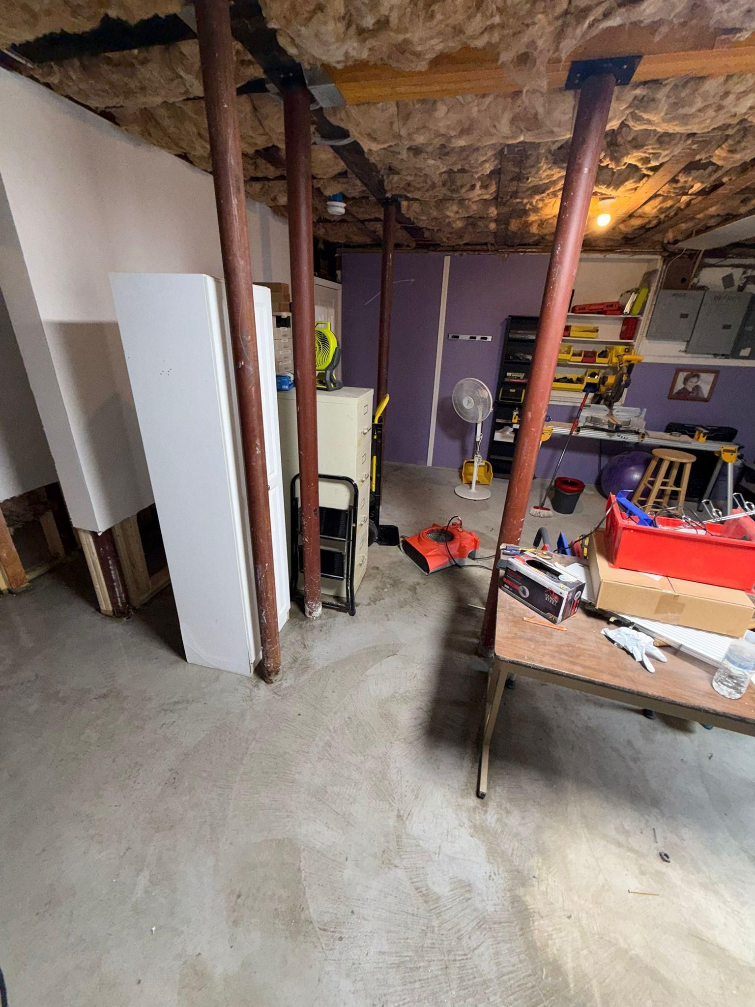 Basement workshop with exposed ceiling, white cabinet, workbench, tools, and scattered equipment.