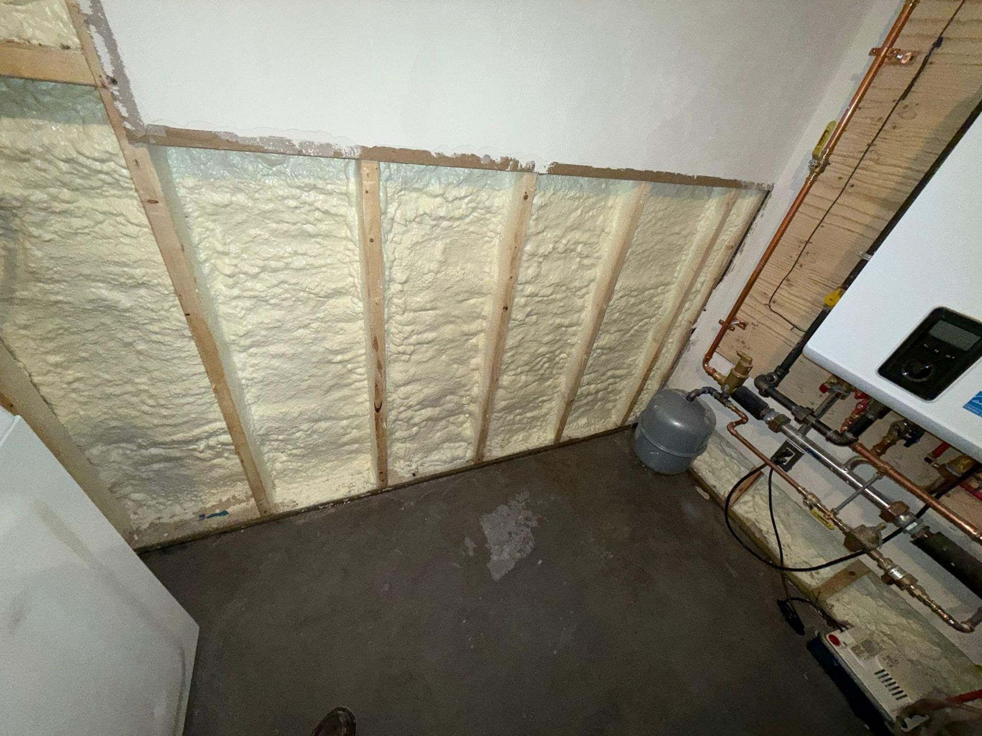 Partially insulated basement wall with exposed studs, concrete floor, and utility piping on the right