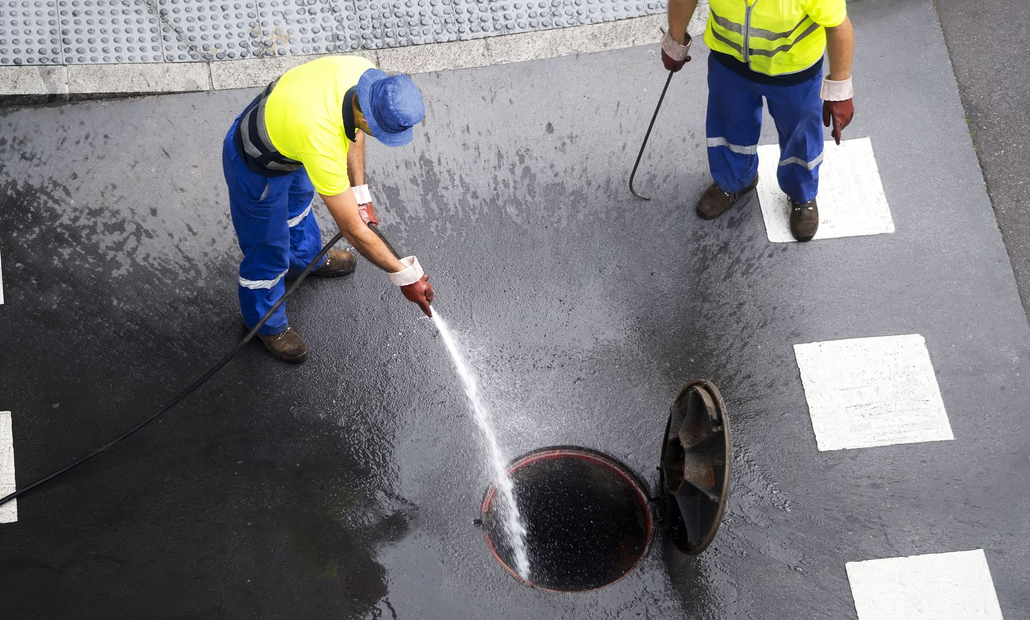 Two road workers in yellow vests cleaning a manhole with a hose on a wet street