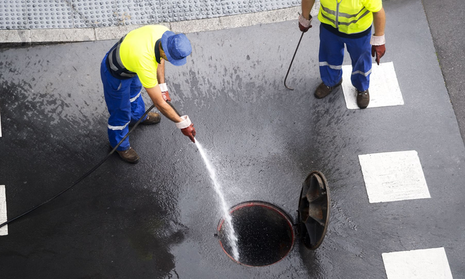 Two road workers in yellow vests cleaning a manhole with a hose on a wet street