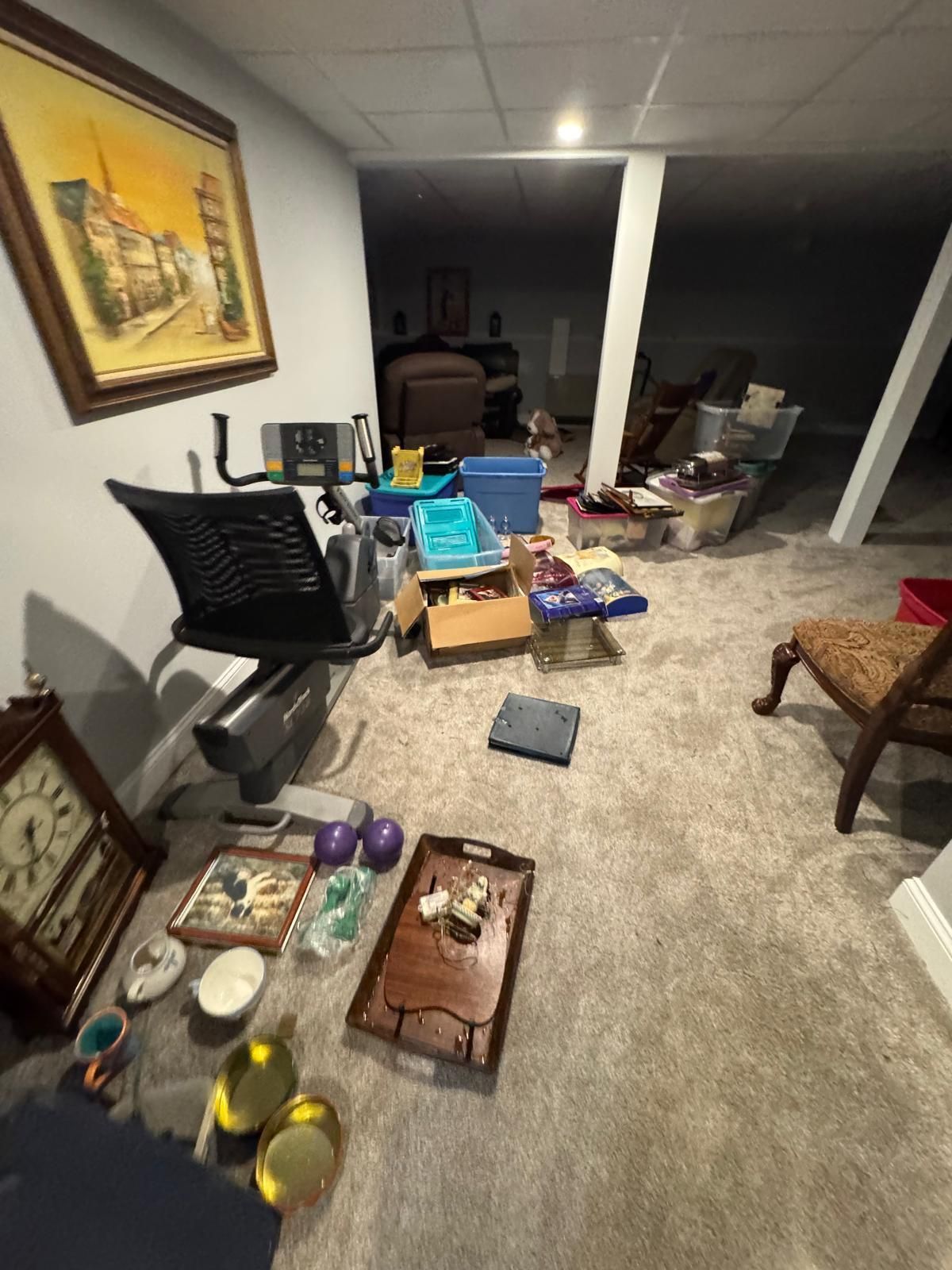 Cluttered basement with boxes, chairs, bins, and stored household items under exposed beams.