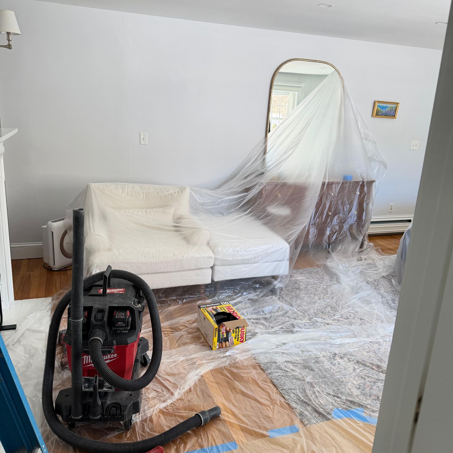 Living room under renovation, with sofa covered in plastic and a vacuum on the floor.