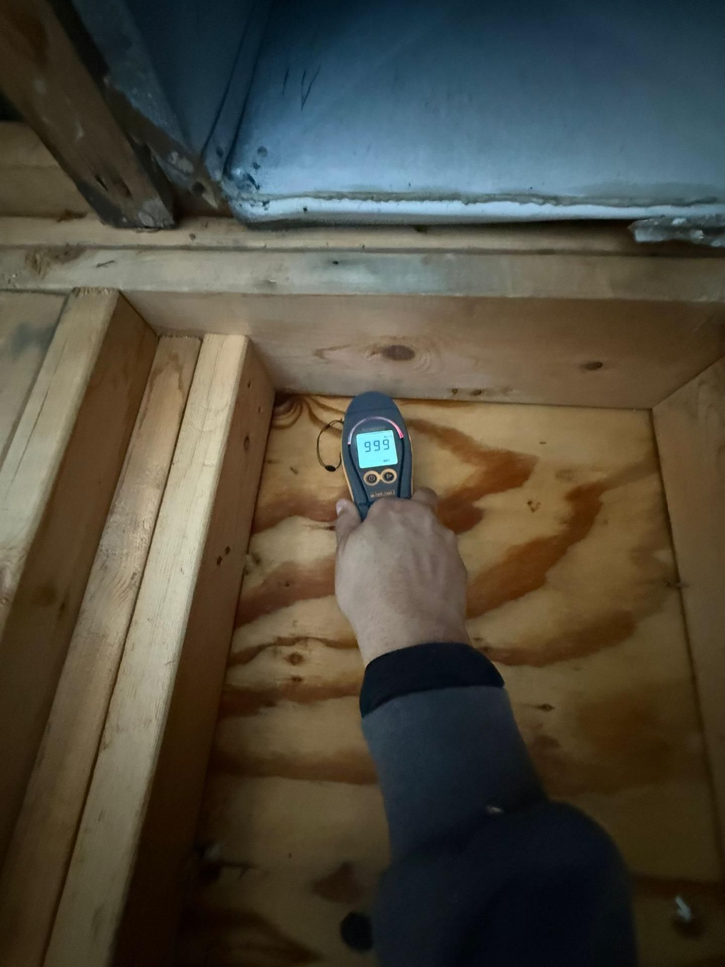 Hand holding a moisture meter against plywood near a wall base in a construction area