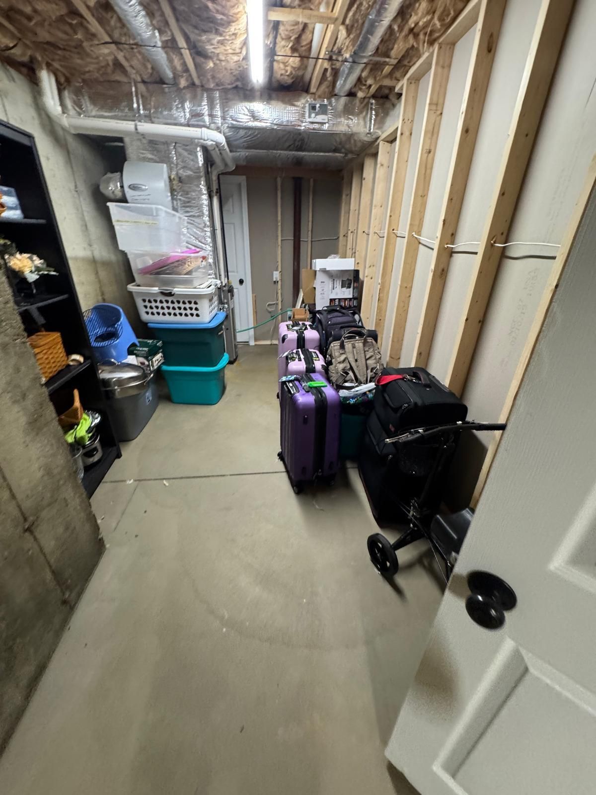Narrow basement hallway with stacked storage bins, luggage, and exposed ceiling pipes.