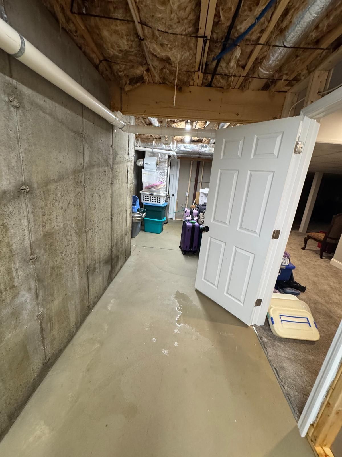 Narrow basement hallway with concrete walls, exposed ceiling, and an open white door leading to a cluttered area