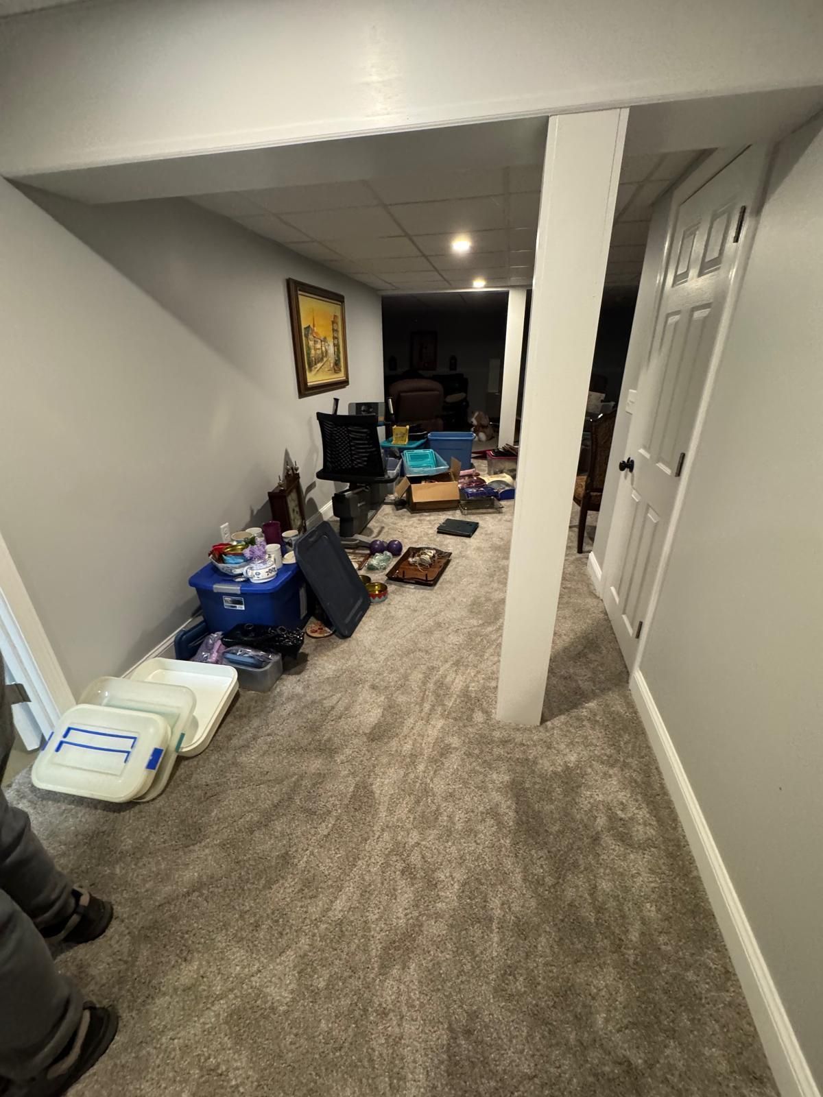 Long basement hallway with cluttered boxes, bags, and furniture along the walls, carpeted floor, and white pillars