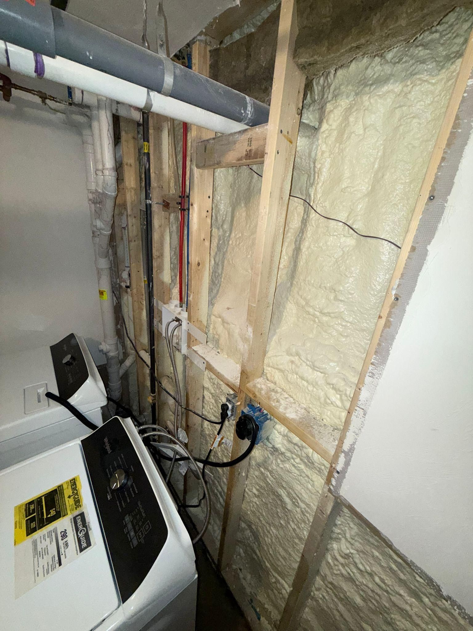 Exposed basement utility wall with pipes, wiring, and insulation beside a white appliance