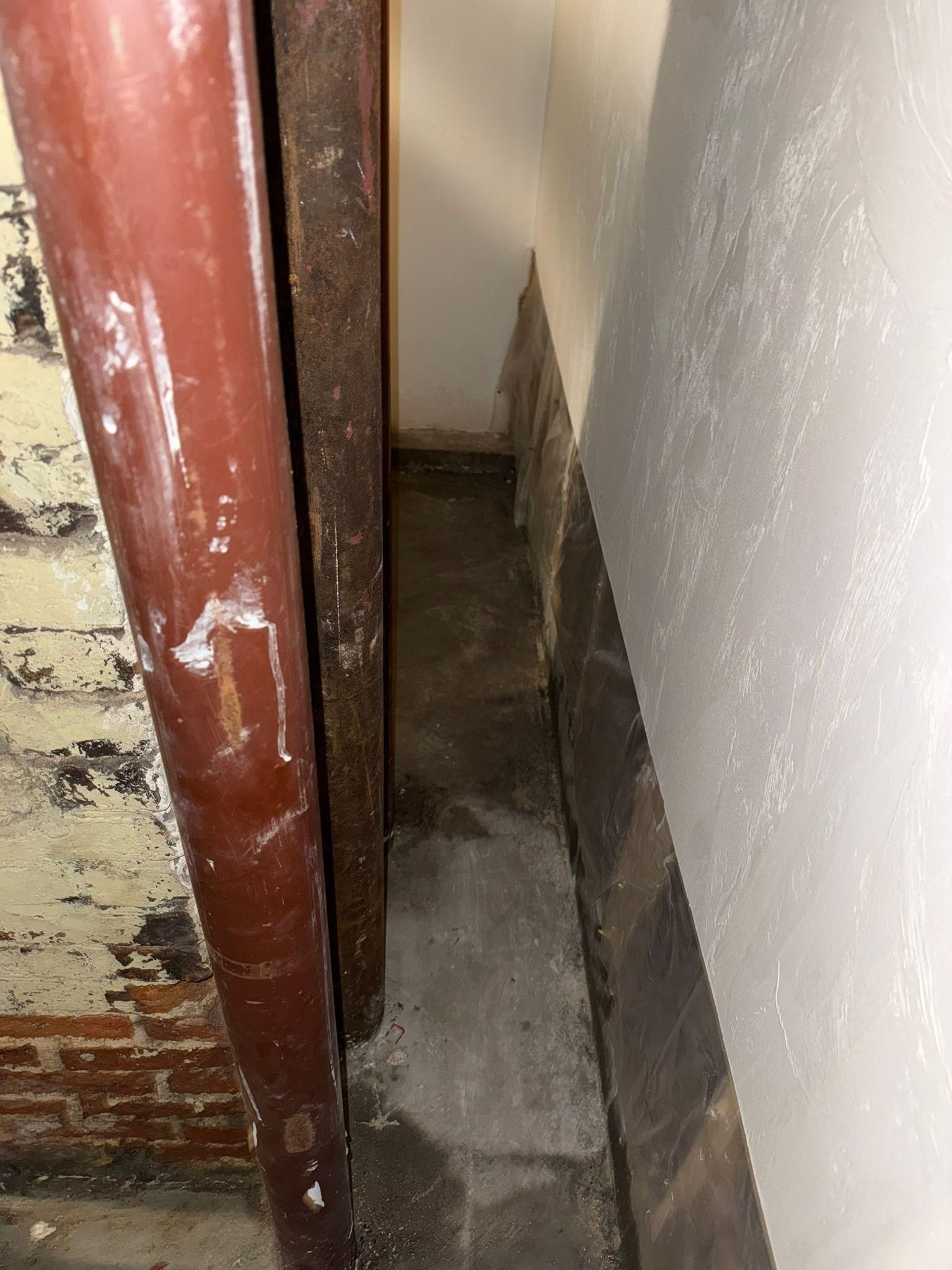 Narrow gap between a red wooden frame and a white wall, showing a rough concrete floor and stained surfaces.