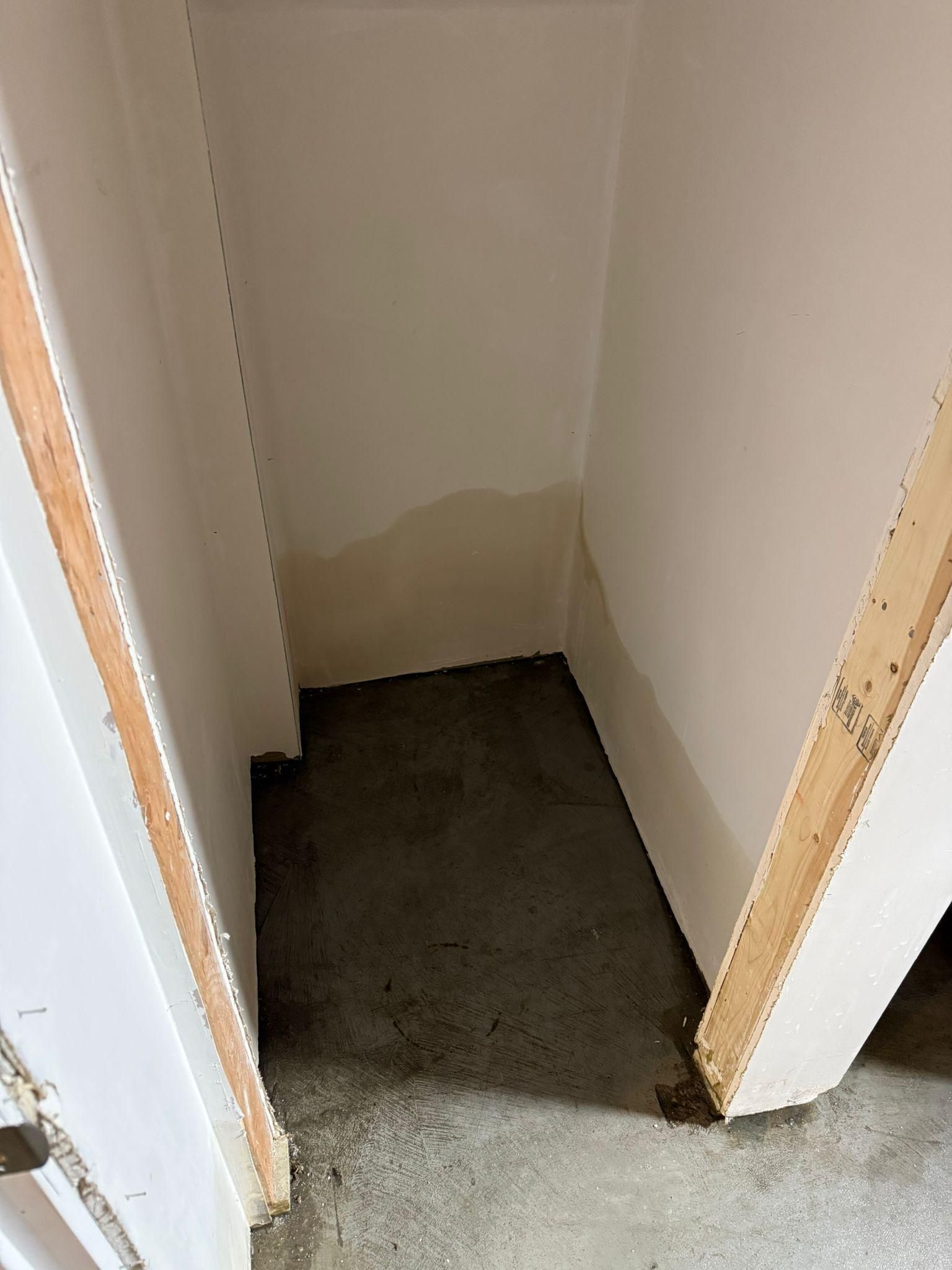 Small unfinished closet with white walls, concrete floor, and exposed wooden door frames.